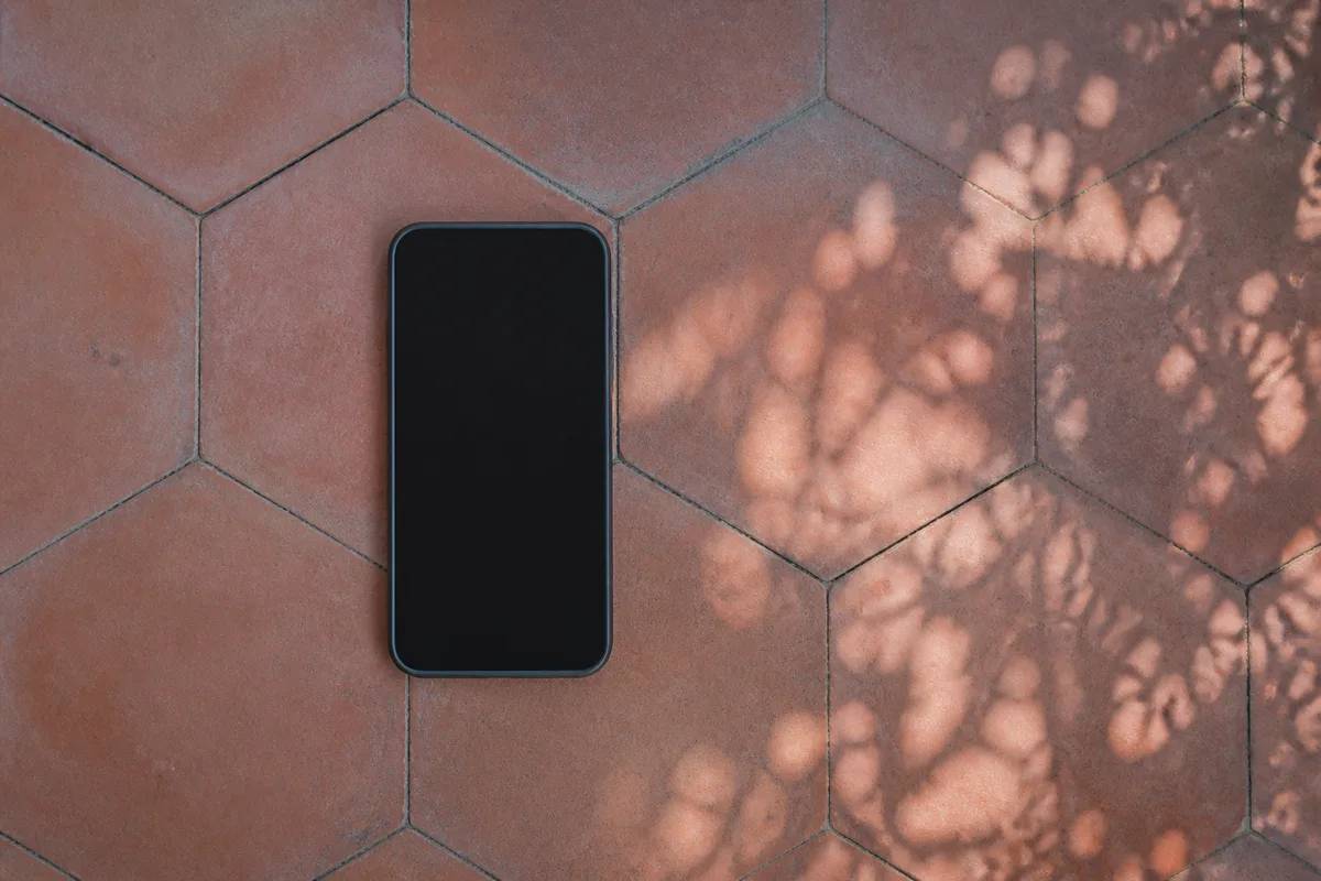 Smartphone à écran éteint posé à plat sur des tomettes en terre cuite à l’ombre d’une véranda
